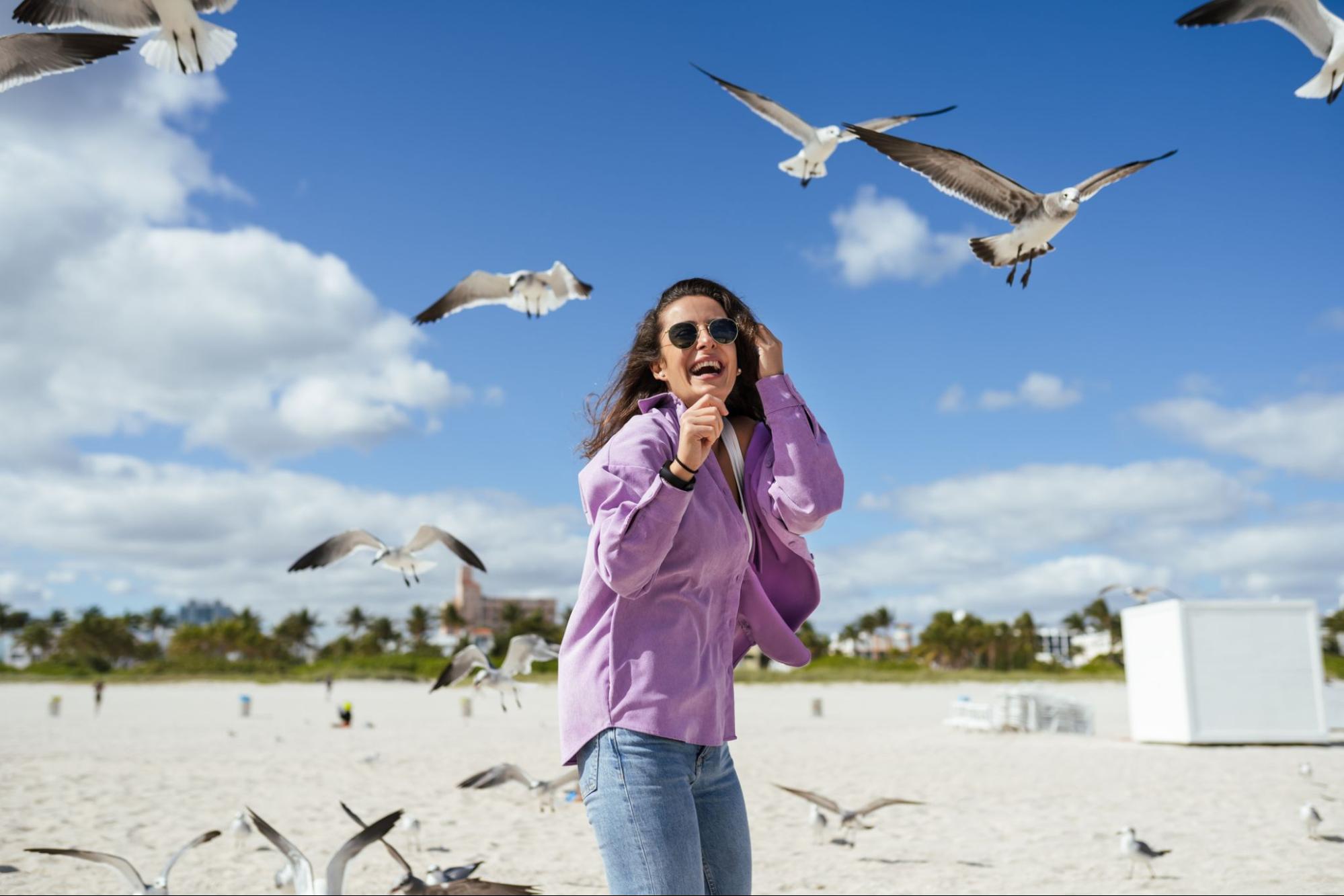 A woman stands on a beach laughing as seagulls fly around her on a sunny day.