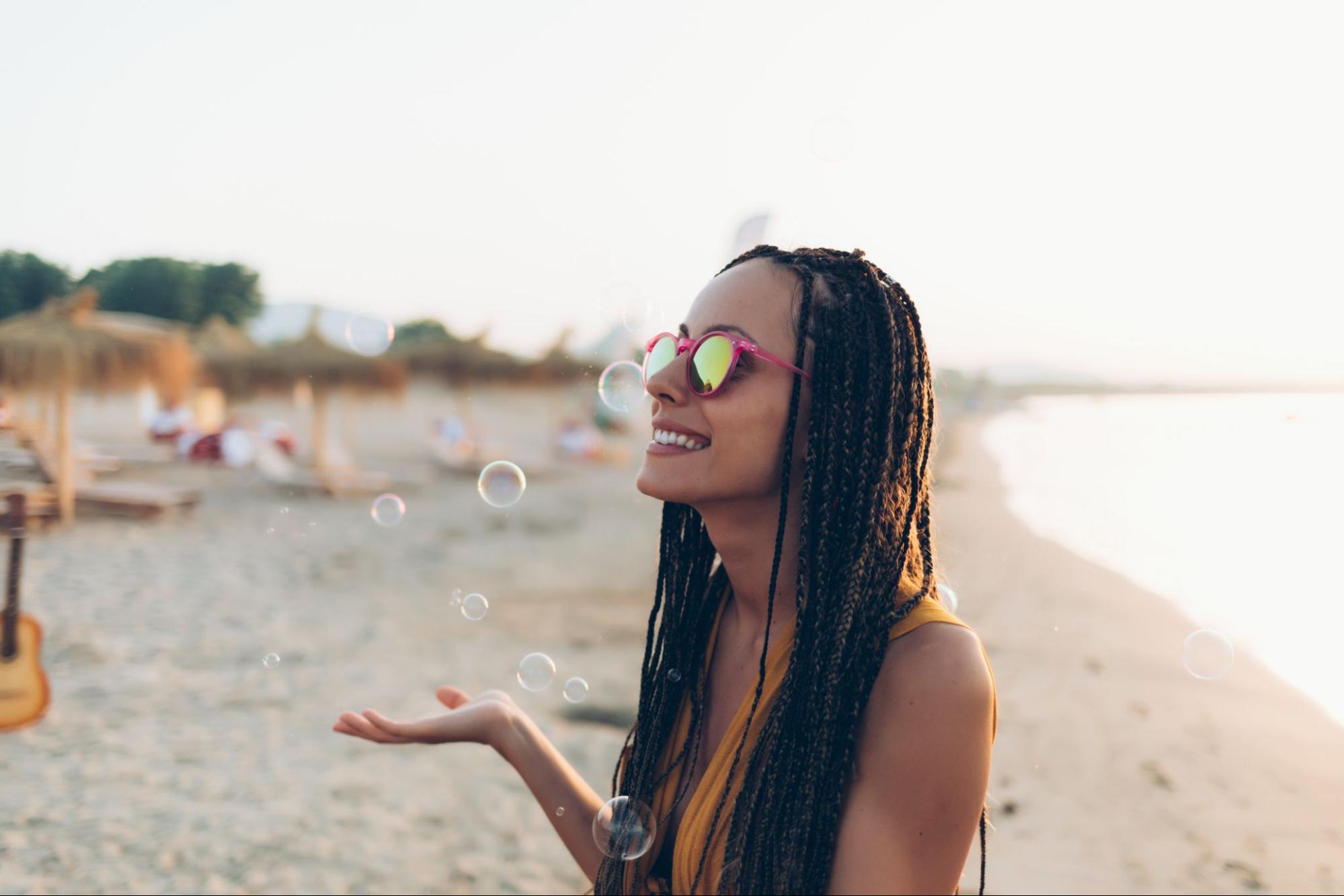 A woman wearing sunglasses smiles and holds up her hands to catch bubbles floating in the air on a beach.